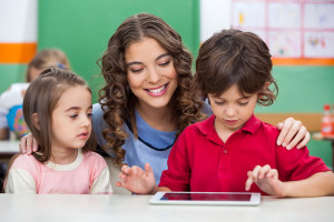 Children using digital tablet with teacher at classroom desk